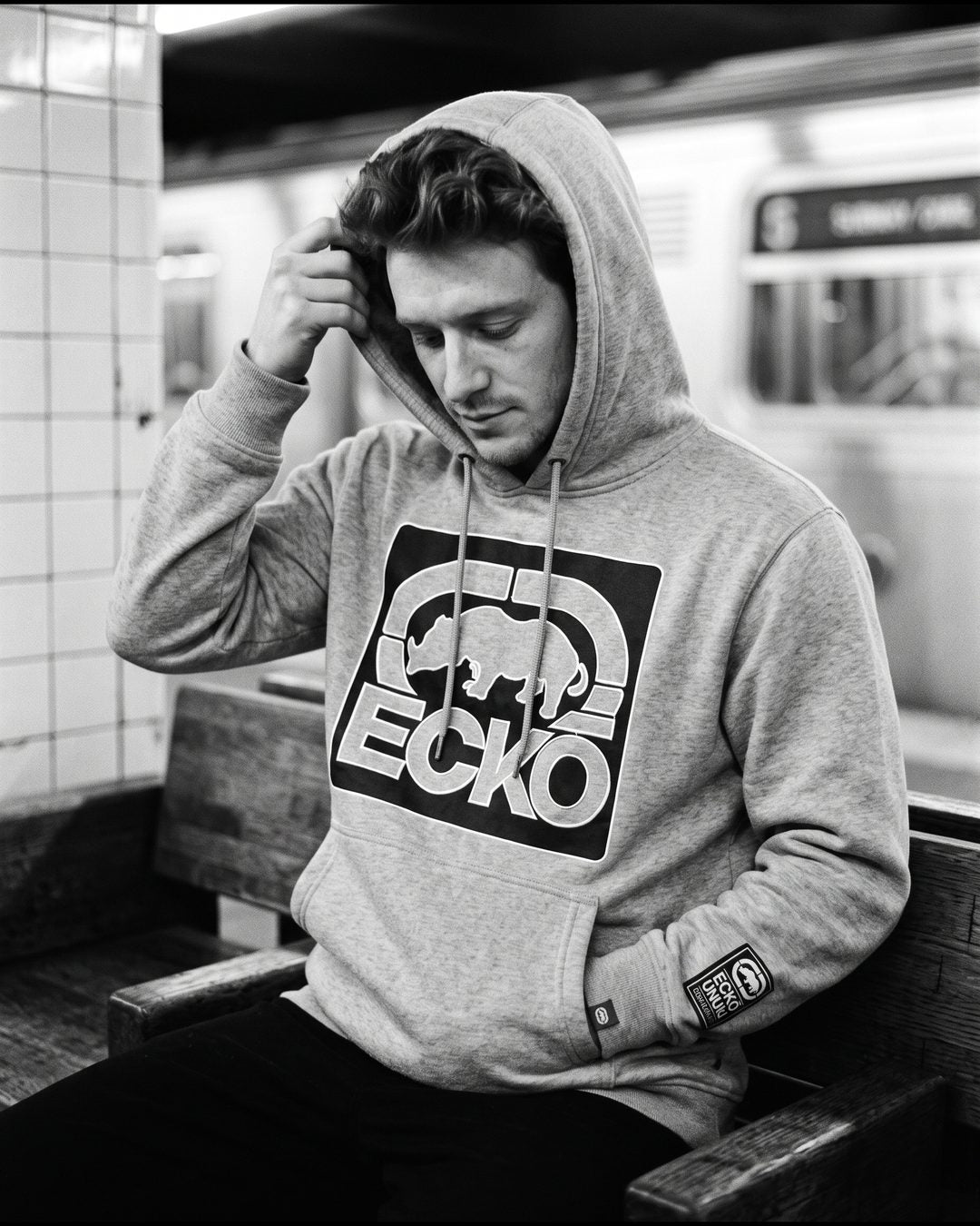 Man pulling up the hood of a grey Ecko Unltd hoodie on a subway platform